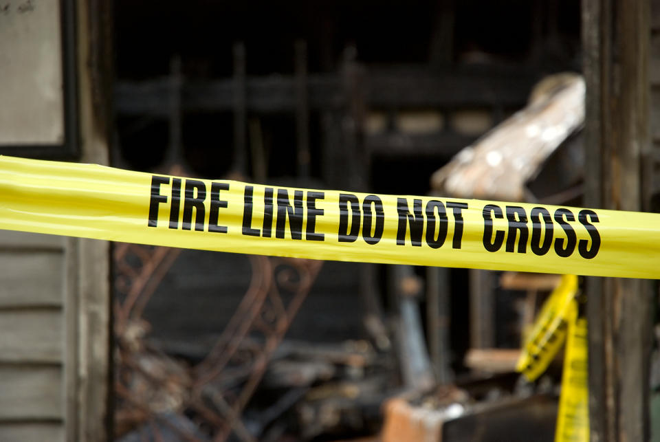 Yellow "Fire Line Do Not Cross" tape blocks access to a burnt building. The structure behind the tape is charred and damaged