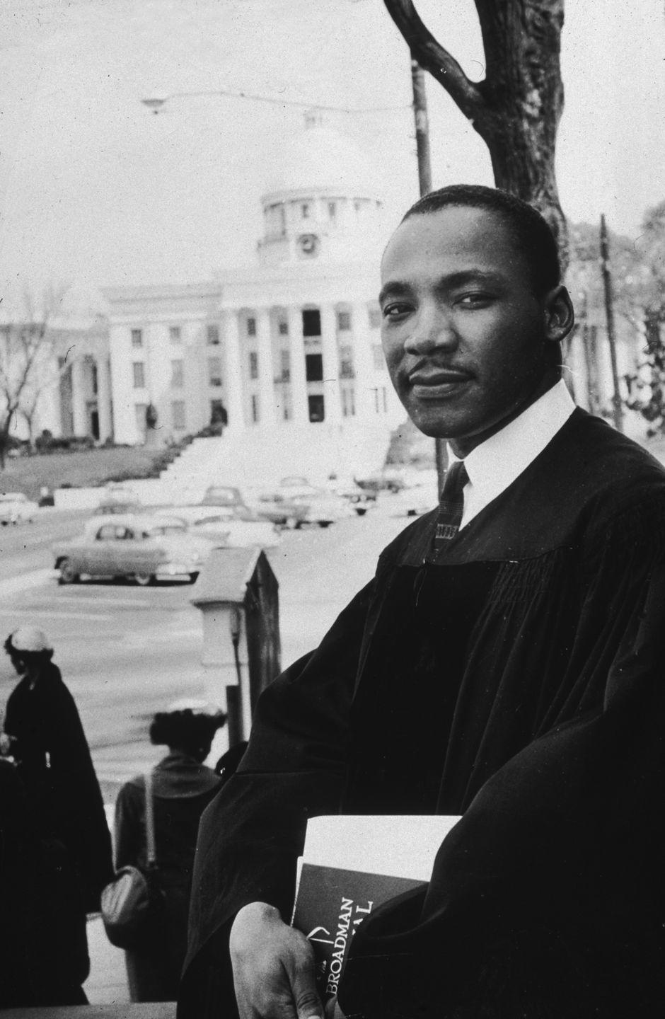 martin luther king jr looks at the camera while standing outside in a pastor robe over a collared shirt and tie, he holds papers in both hands in front of him, behind him is a street scene and a large white building