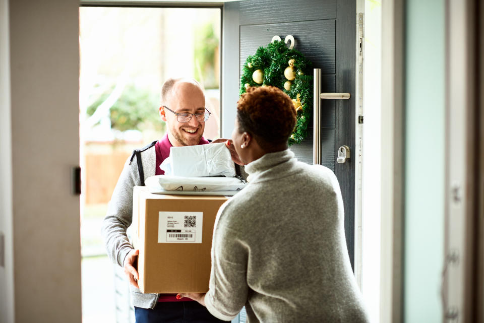A man in glasses delivers packages to a woman at her front door adorned with a wreath. The woman wears a sweater and smiles while receiving the packages