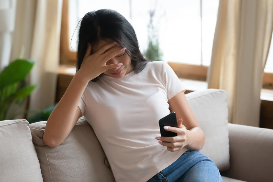 Frustrated asian woman sit on couch holds smart phone read message unpleasant sms cover face with hands feels upset, crying. Personal problems, life troubles, break up with boyfriend, bad news concept