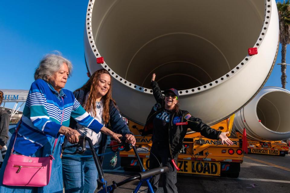 A woman with a walker is assisted by another as a third woman poses for photo near a large metal cylinder.