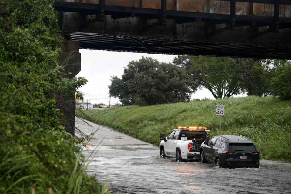 A tow truck pulls an SUV through a flooded underpass under a railroad bridge after heavy rains. Speed limit sign shows 40 mph. Trees and vegetation are visible