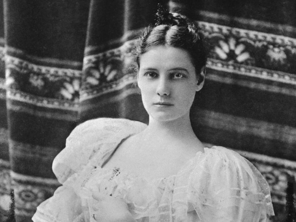 Nellie bly sits in a chair in a puffy-sleeved dress with her hair up and a patterned blanket in the background