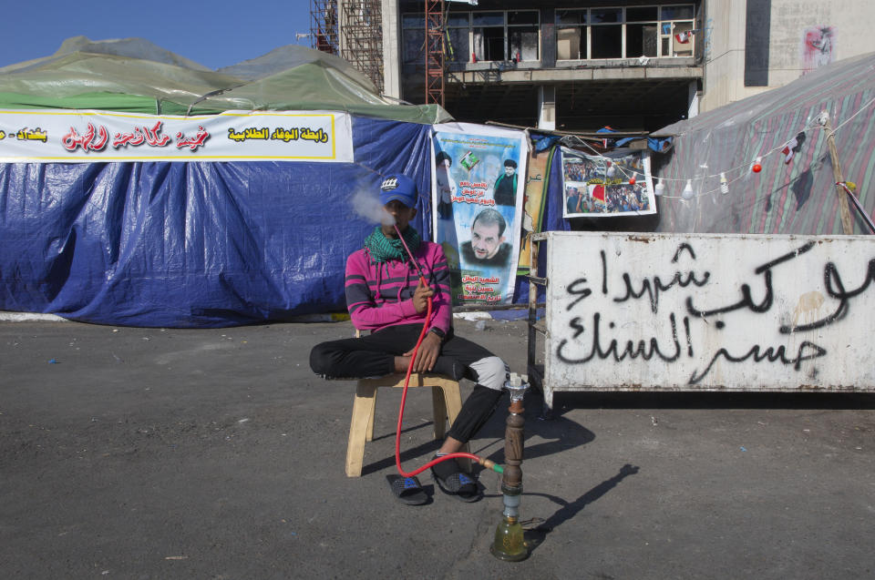 In this Thursday, Dec. 19, 2019, photo, a youth protester rests and smokes a water pipe next to Arabic that reads "the convoy of the Al-Sanak bridge Martyrs," during the ongoing protests in Tahrir square, Baghdad, Iraq. Tahrir Square has emerged as a focus of the protests, with protesters camped out in dozens of tents. Dozens of people took part in the simple opening of the sculpture exhibition. (AP Photo/Nasser Nasser)