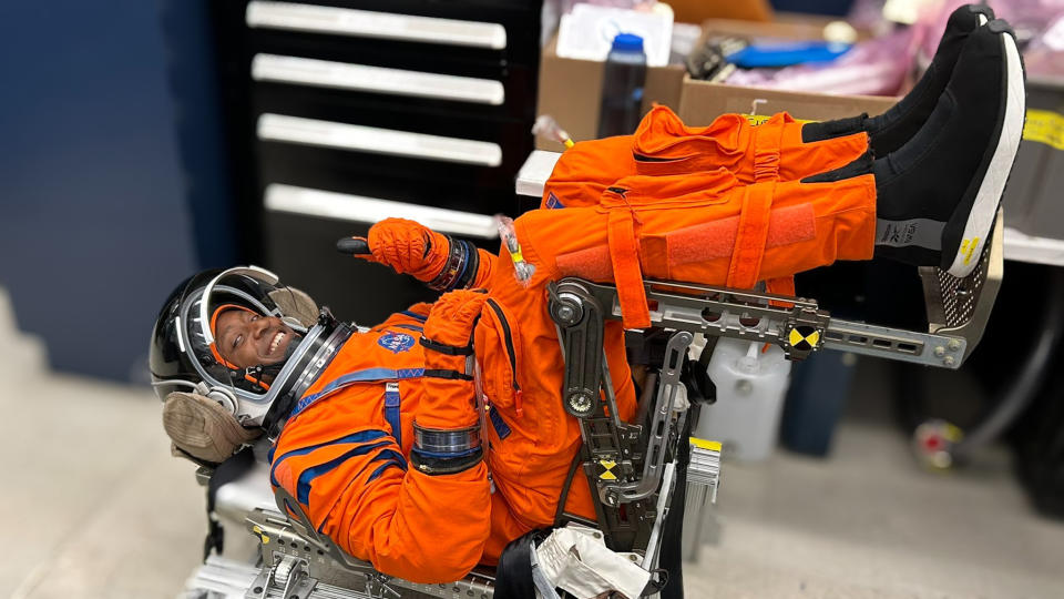  An astronaut giving a thumbs-up inside a spacesuit, while lying on his back near a filing cabinet. 