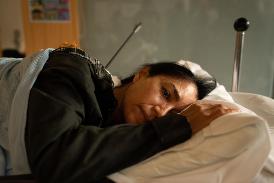 Niamh Kirby (Seeta Indrani) lies on her front on a stretcher being wheeled through a hospital corridor, still wearing her clothes and her outdoor coat, with an arrow sticking out of her back