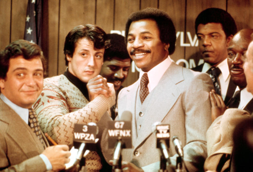 Sylvester Stallone in a patterned sweater and Carl Weathers in a grey suit stand together at a press event with microphones