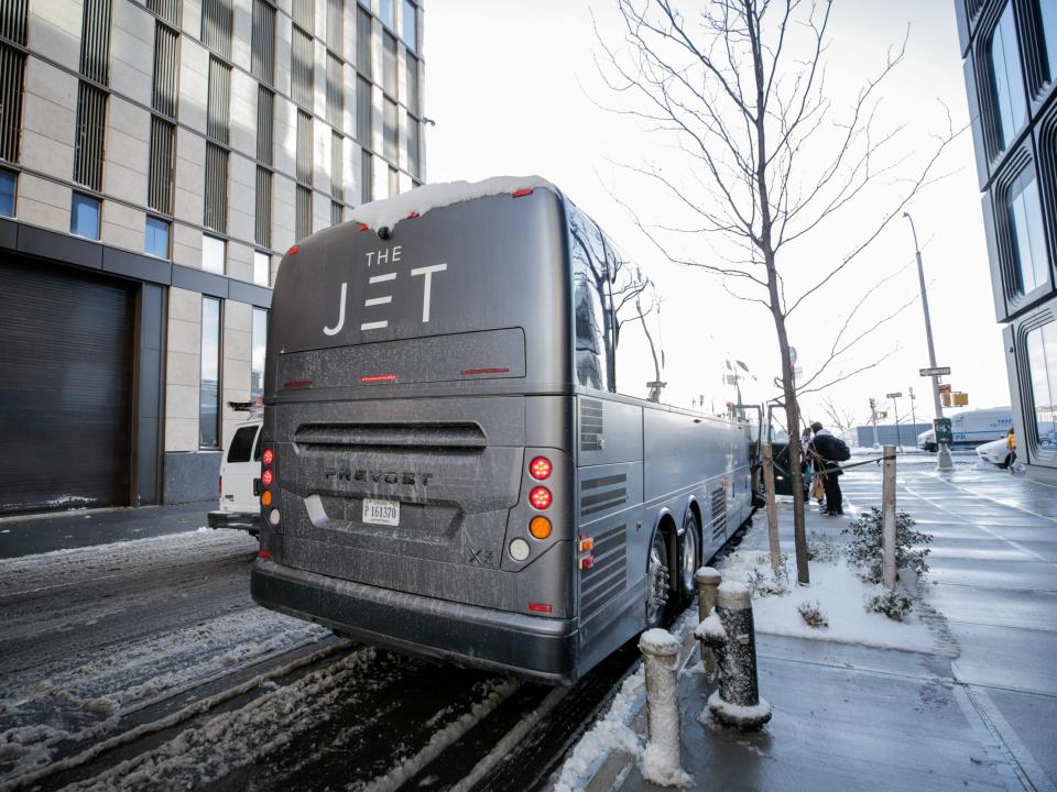 A matte black bus that reads "The Jet" on the side. Passengers with bags are boarding the bus or putting their bags away into the lower storage compartment.