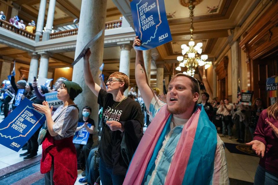 Kristopher Carlock listens to speakers with a transgender pride flag draped around his shoulders Saturday, April 1, 2023, during a Rally to Protect Trans Youth organized by the ACLU of Indiana at the Statehouse in Indianapolis.