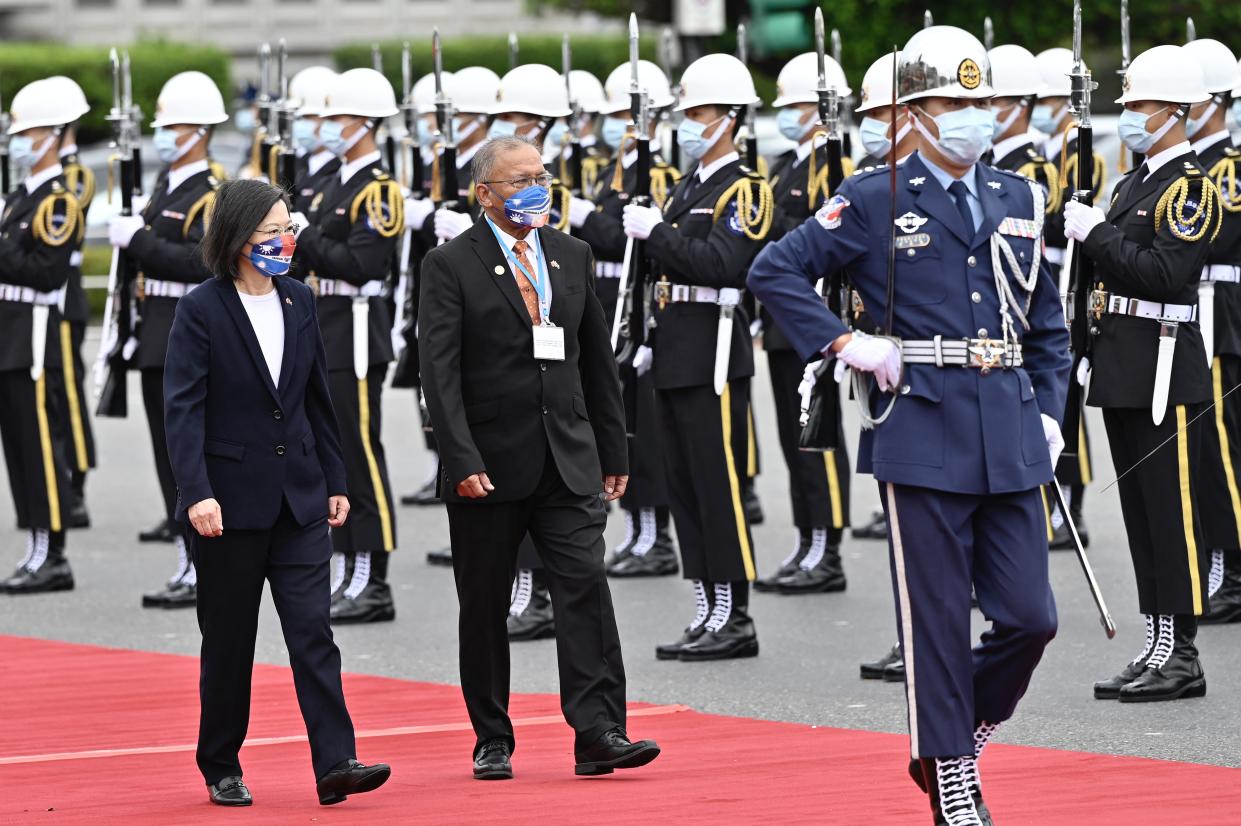 Taiwan President Tsai Ing-wen and Marshall Islands President David Kabua walk along a red carpet while a few dozen troops with white helmets, blue face masks, and rifles with bayonets stand at attention.