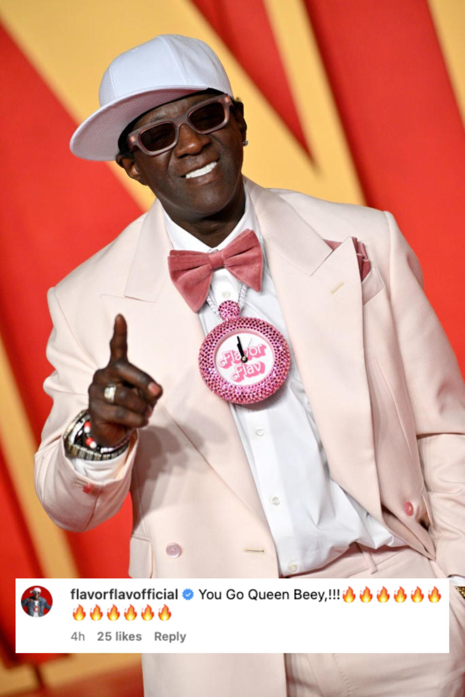 Flavor Flav in a light suit, bow tie, and hat, flashing a smile and pointing at the camera