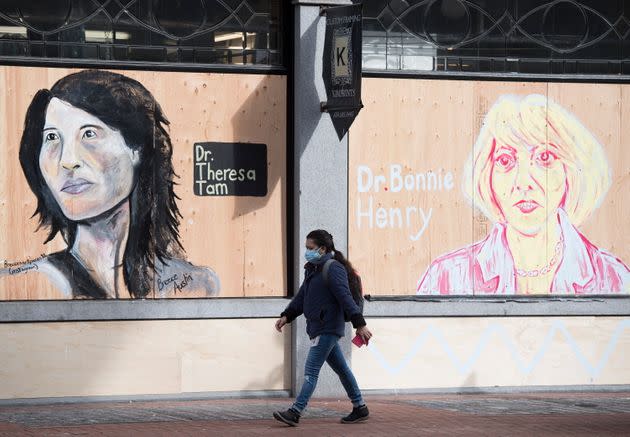 A woman wearing a protective face mask walks past portraits of Dr. Theresa Tam and Dr. Bonnie Henry on a boarded up business in downtown Vancouver, B.C. on April 1, 2020.