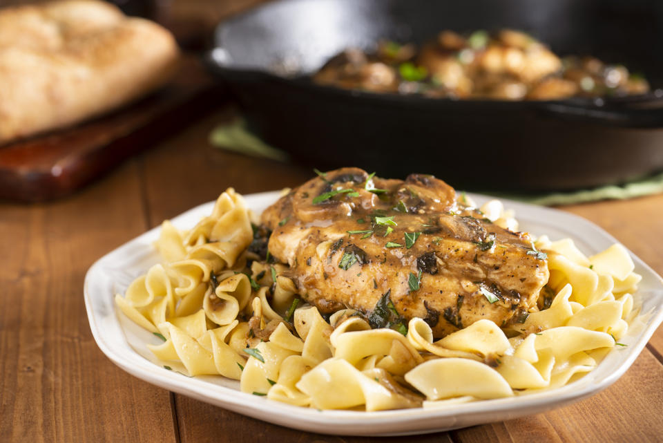 A plate of cooked egg noodles topped with a piece of chicken and mushroom sauce, with a bread loaf and a pan in the background