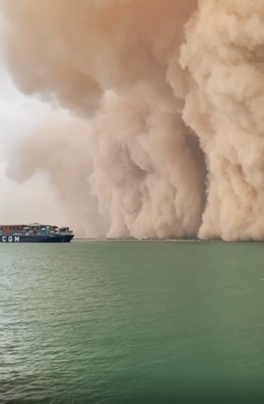 CGM cargo ship sailing near billowing clouds of smoke or sand over the ocean;  dramatic environmental scene