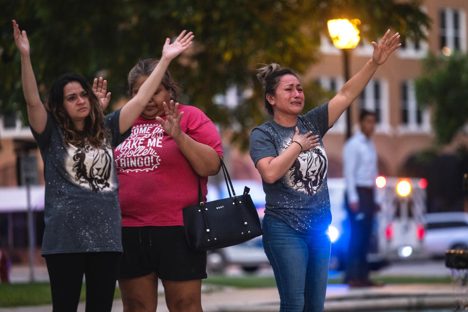 Three people stand near a street holding their hands in the air in prayer.