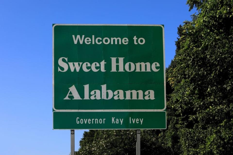 Road sign reads "Welcome to Sweet Home Alabama" with "Governor Kay Ivey" below, set against a backdrop of trees and a clear sky