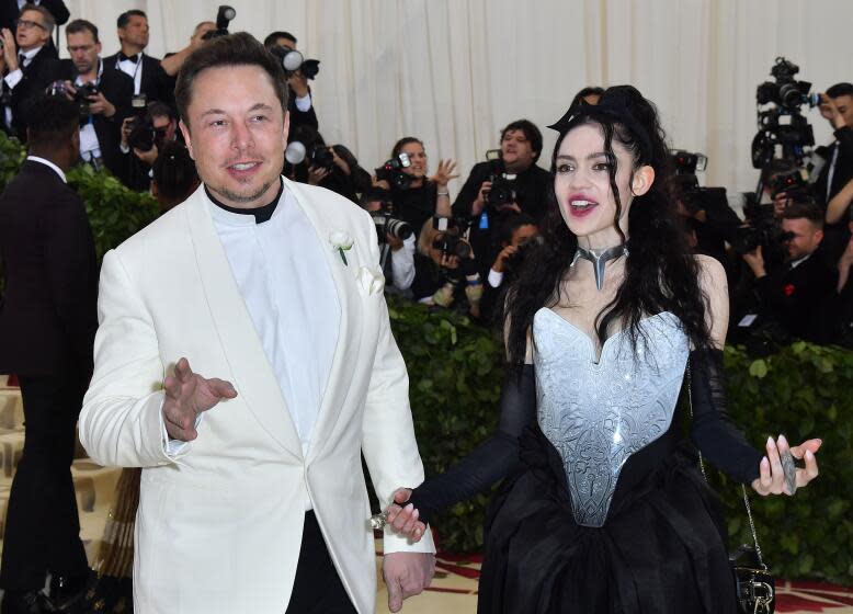 Elon Musk and Grimes speak while wearing formal attire in front of a crowd of photographers.