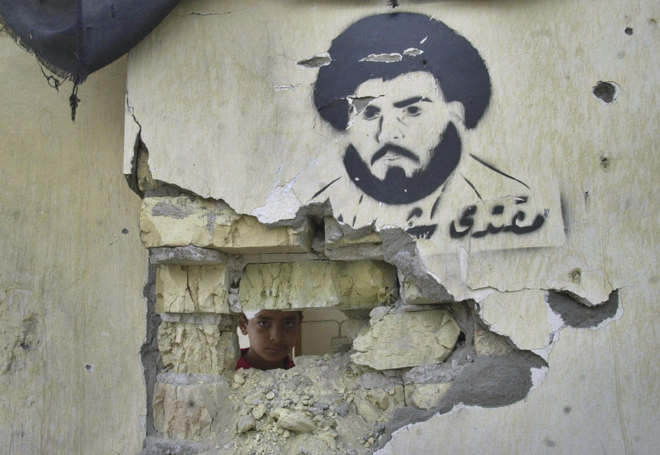 FILE - An Iraqi boy peers through a destroyed wall with a painting of militant Shiite cleric Moqtada al-Sadr in the Sadr City district in Baghdad, Iraq, Monday, May 10, 2004. Al-Sadr is a populist cleric, who emerged as a symbol of resistance against the U.S. occupation of Iraq after the 2003 invasion. (AP Photo/Khalid Mohammed, File)
