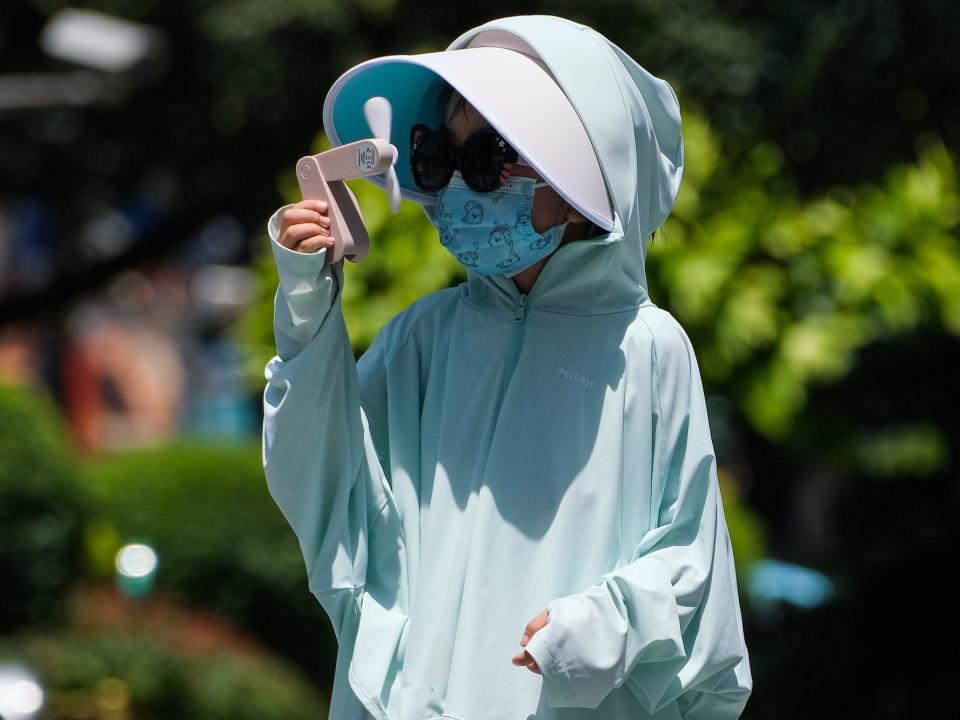 A child wearing a mask, a visor and a light-protective hoody holds an electric fan to their face.