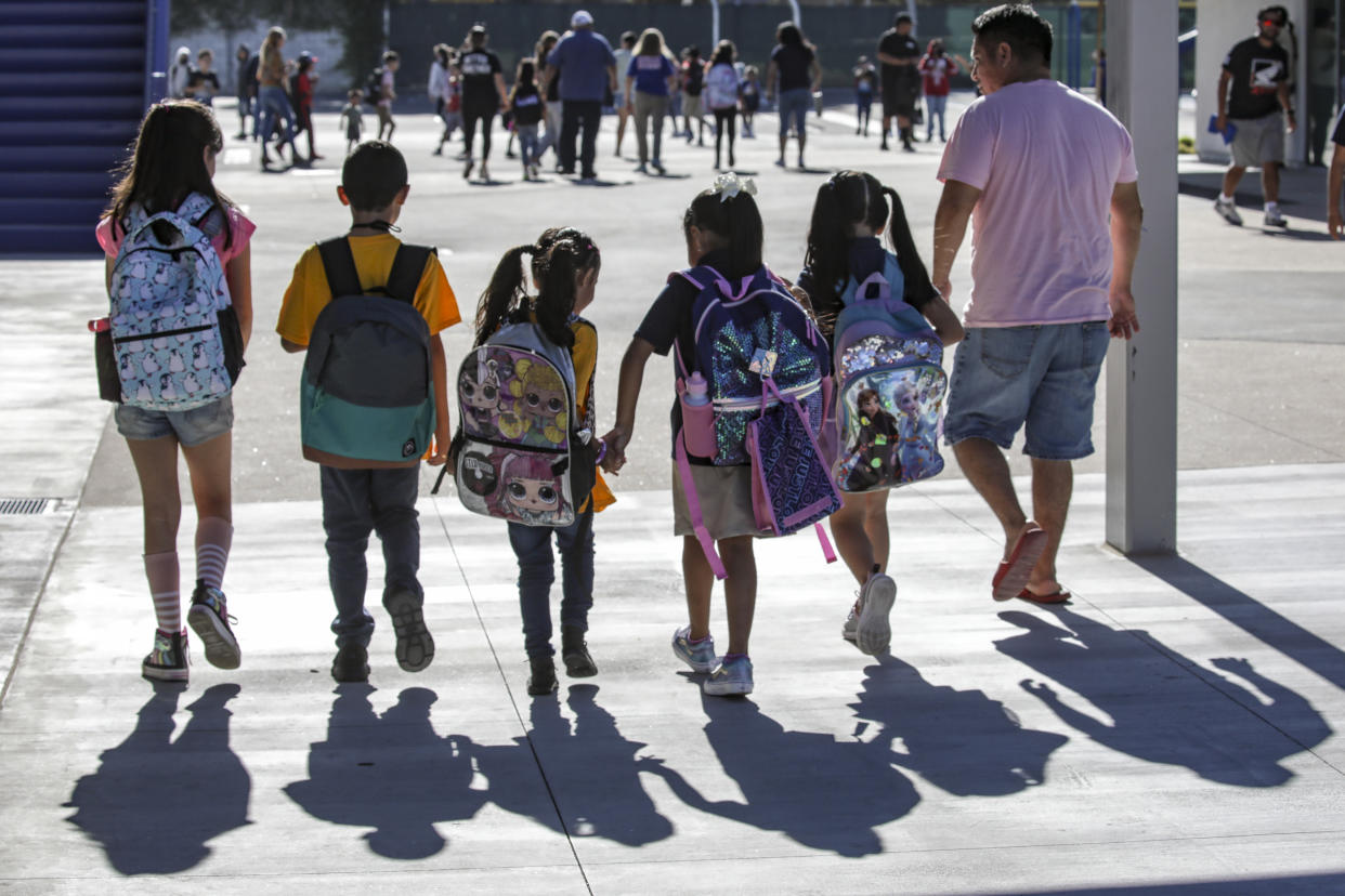 Five students with colorful backpacks walk toward a school.