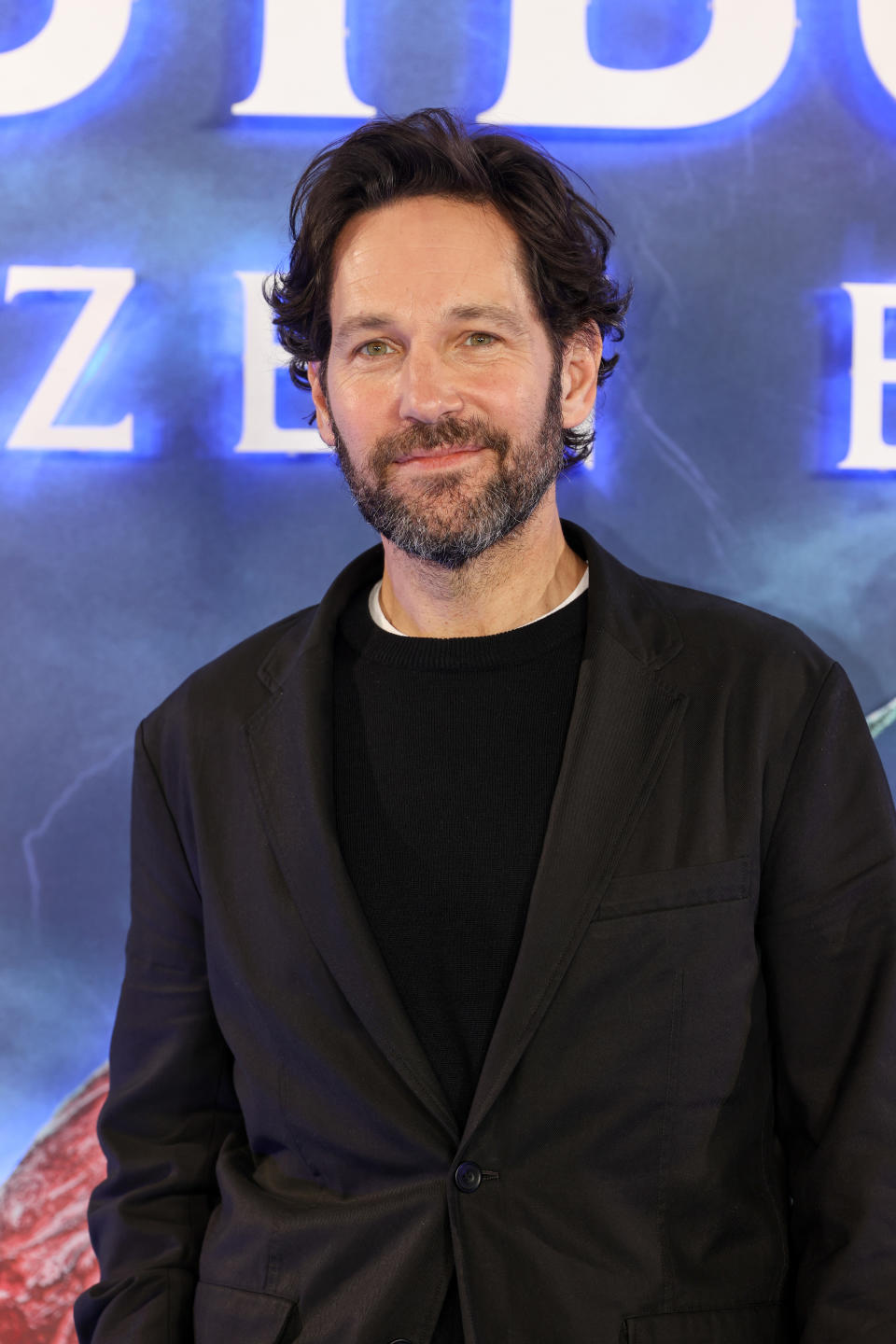 Paul Rudd in a black suit stands smiling at a "Ant-Man and the Wasp: Quantumania" event