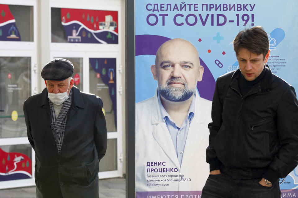 Two men, one of them wearing a face mask, stand near a vaccination point decorated with the poster showing a portrait of Dr. Denis Protsenko and words reading "Get vaccinated against covid-19!!" at VDNKh, The Exhibition of Achievements of National Economy in Moscow, Russia, Friday, April 2, 2021. Moscow is one of the few places in the world where one can get vaccinated against COVID-19 within hours of deciding to do so. Free doses of the domestically developed Sputnik V shot are readily available for anyone 18 or older at more than 200 vaccination points in state and private clinics, shopping malls, food courts, hospitals and even a theater. (AP Photo/Alexander Zemlianichenko)