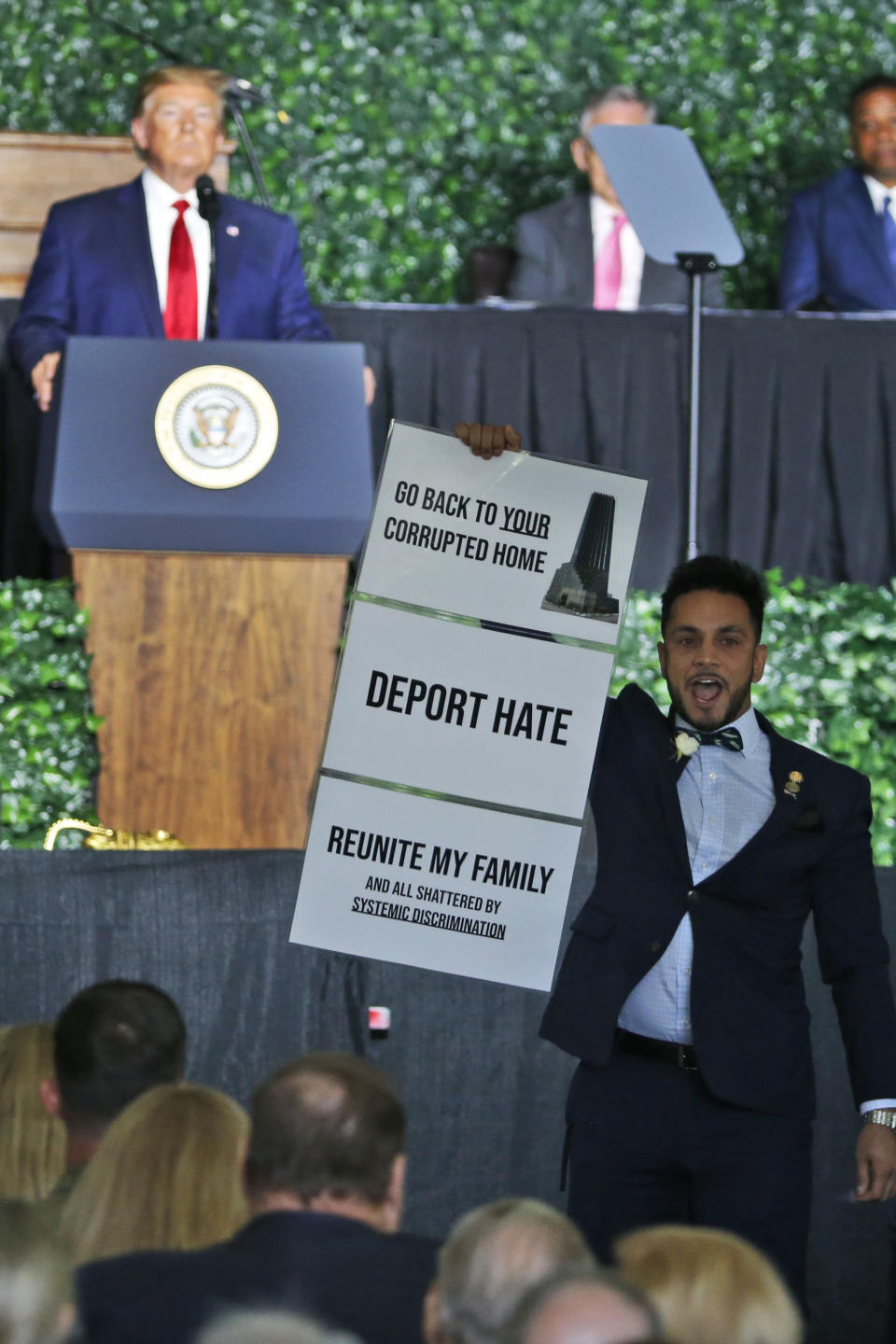 ADDS NAME OF SAMIRAH - President Donald Trump waits as Virginia Delegate Ibraheem S. Samirah, D-Fairfax, holds a sign and interrupts a speech by Trump as he addresses a commemorative meeting of the Virginia General Assembly at Jamestown Settlement on the 400th anniversary of the meeting of the original House of Burgess in Jamestown, Va., Tuesday, July 30, 2019. (AP Photo/Steve Helber)