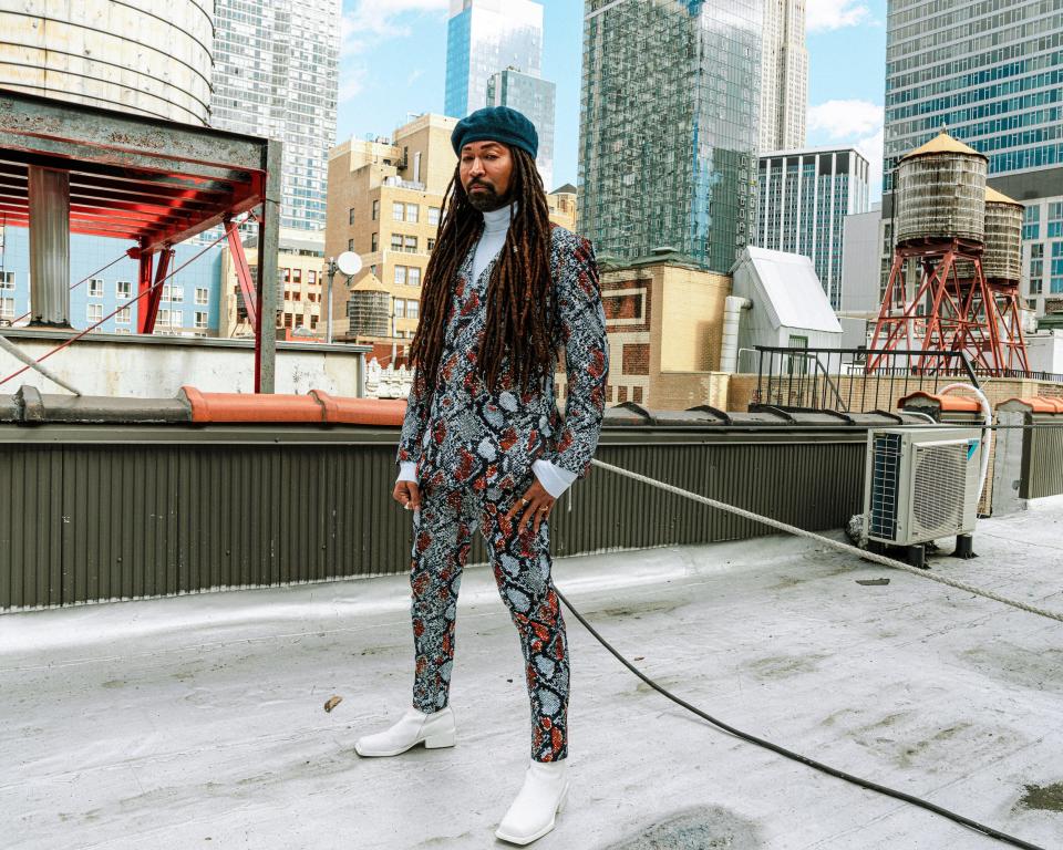 Ty Hunter stands on a rooftop while wearing a black and red snakeskin blazer over a white turtleneck with white boots. A cityscape of New York is in the background.