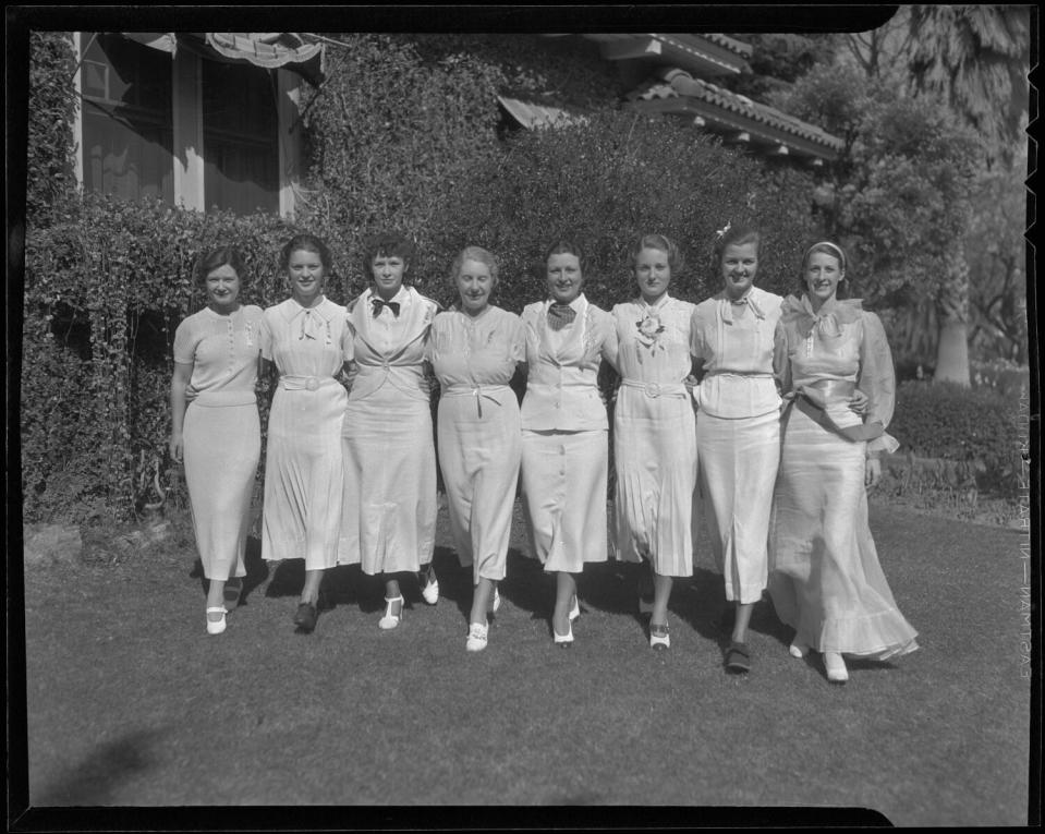 Eight women wearing light-color dresses pose for a black-and-white photograph on a lawn in lush surroundings.