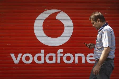 A man checks his mobile phone as he walks past a shop displaying the Vodafone logo on its shutter in Mumbai January 15, 2014. REUTERS/Danish Siddiqui/Files