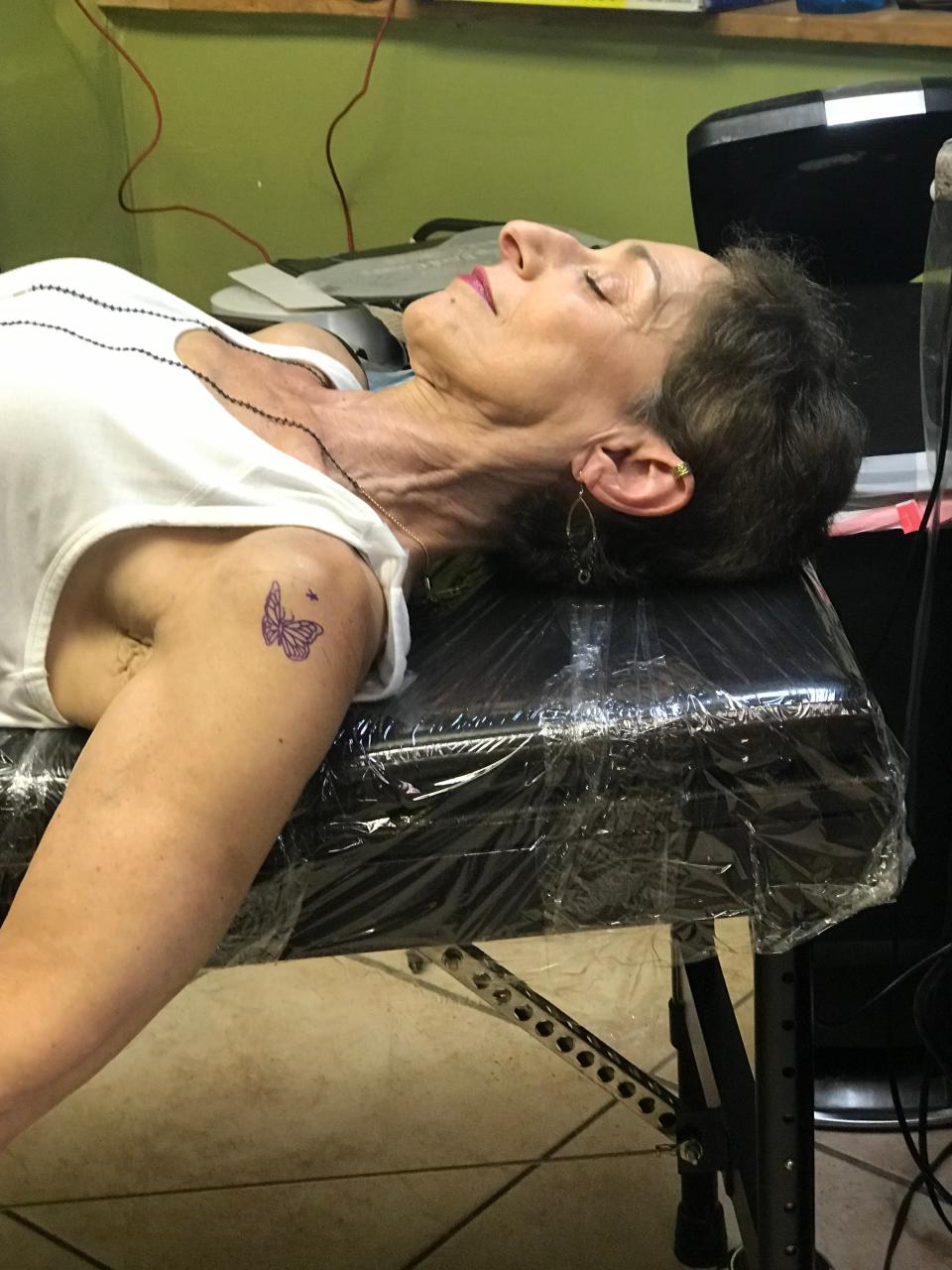 The author in a tattoo shop lying down on a tattooing bench with a stencil of a butterfly tattoo her upper arm waiting to be tattooed