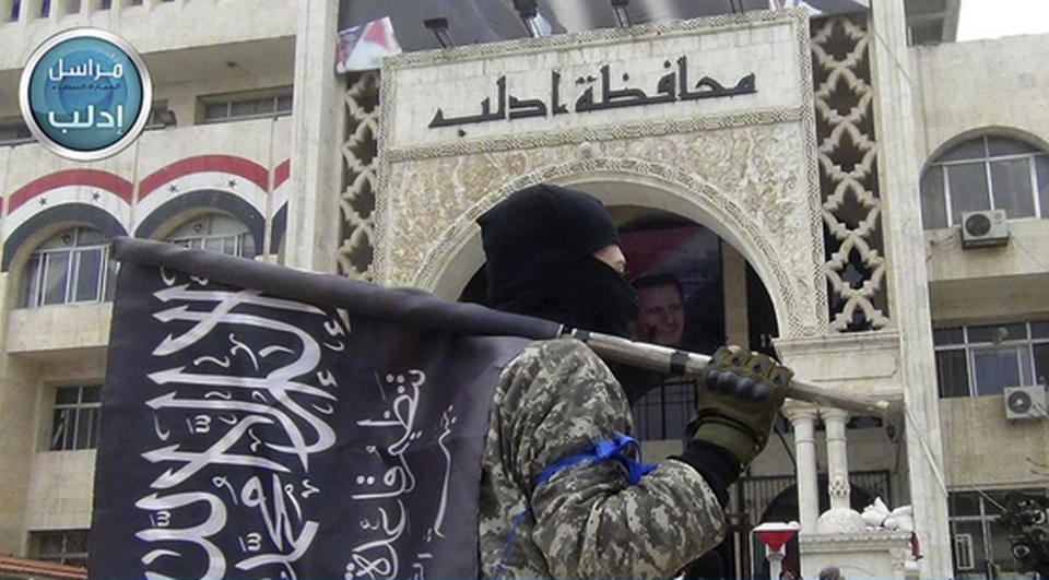 FILE - In this file photo posted on the Twitter page of Syria's al-Qaida-linked Nusra Front on March 28, 2015, a fighter from Syria's al-Qaida-linked Nusra Front holds his group flag as he stands in front of the governor building in Idlib province, north Syria. Intermingled among 3 million civilians under siege in the Syrian government's assault on the last opposition stronghold are tens of thousands of al-Qaida-linked fighters and other militants who came from around the world to take part in the country's civil war. (Al-Nusra Front Twitter page via AP, File)