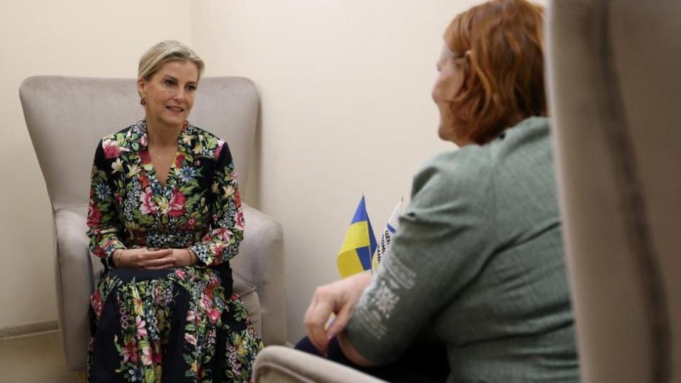 Sophie sits on an armchair, speaking to a woman with red hair whose face cannot be seen by the camera