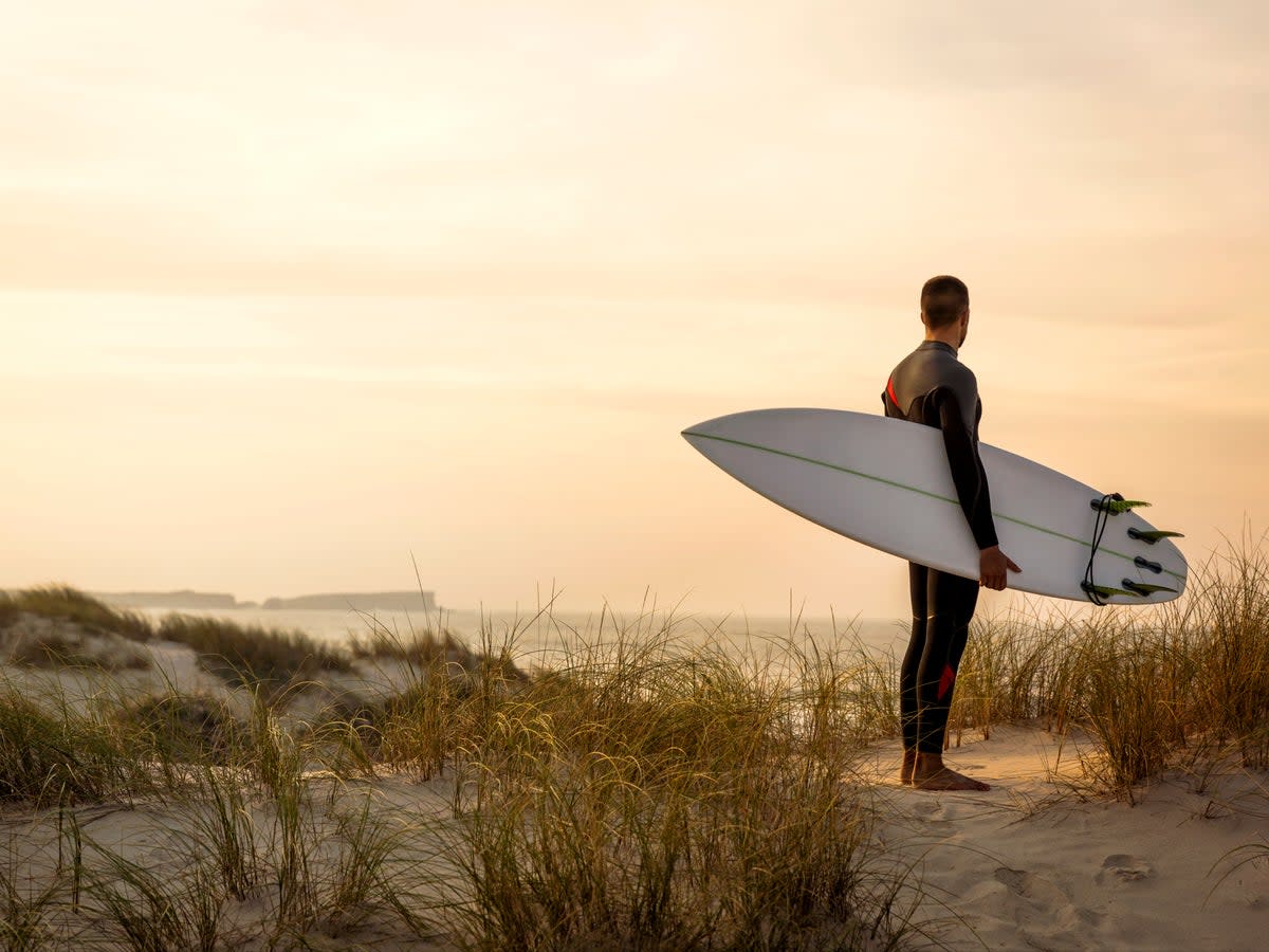 A surfer with his surfboard at the dunes looking to the waves (Getty Images/iStockphoto)