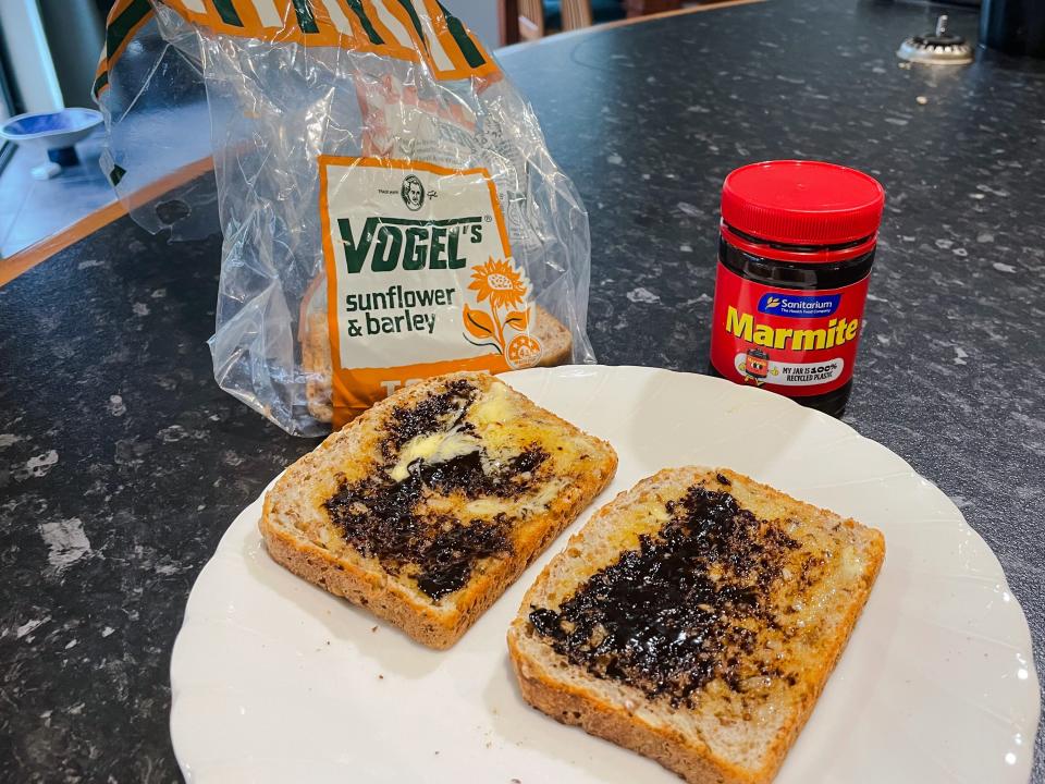 Slices of bread on a white plate with a jar, with a red label that says "marmite" on it in yellow lettering next to it. The plate sits on a black countertop next to a nearly empty clear plastic bag of bread.