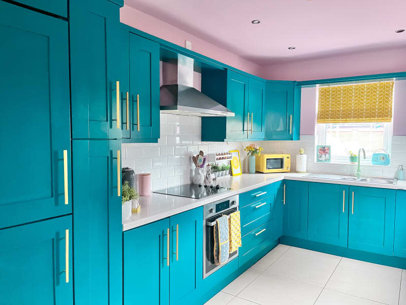 Kitchen after renovation/painting. Bright turquoise cabinets and drawers, pink ceiling, white counters, white subway tile backsplash, bright brass/gold hardware, yellow and pink accents