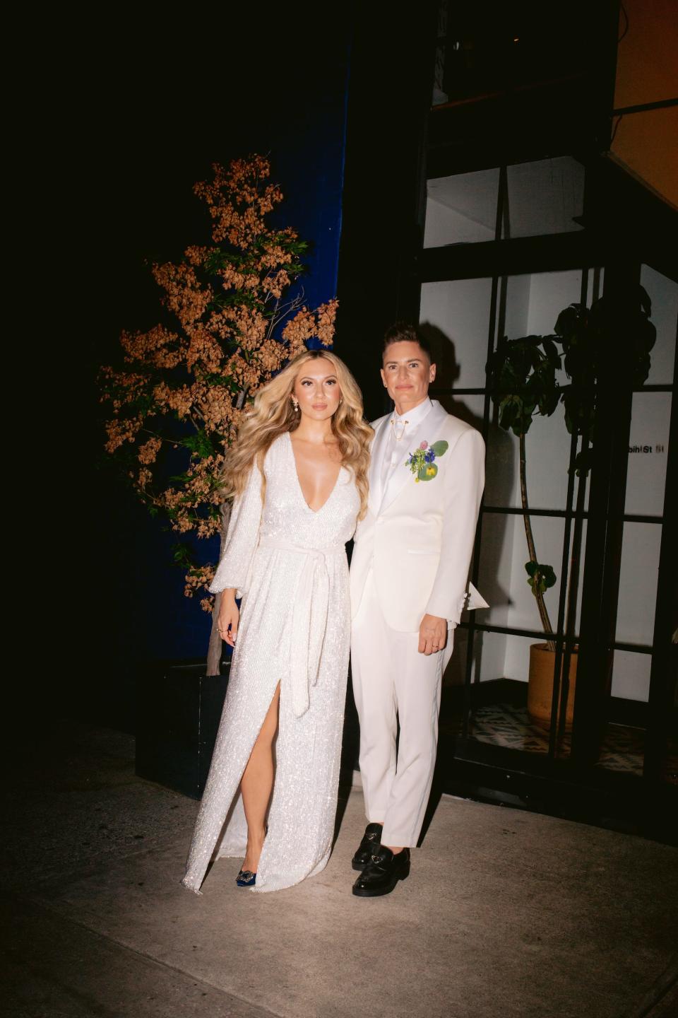 A bride in a white long sleeve dress and a bride in a white suit pose for a photo.