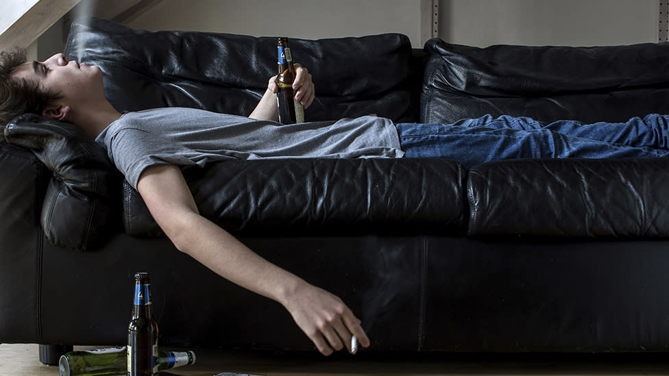 Un joven echado en un sofá fumando y bebiendo cerveza