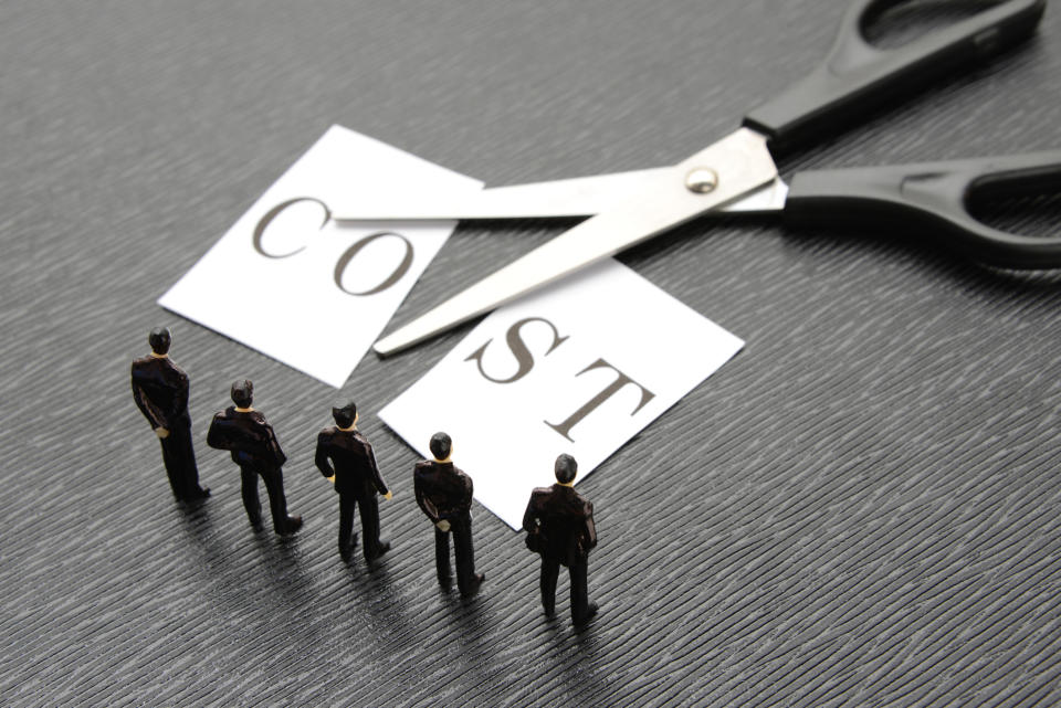 Scissors cutting a paper with the word Cost printed on it, as tiny model businesspeople look on