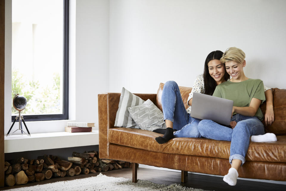 Two people sitting closely on a couch, smiling and looking at a laptop together in a cozy living room