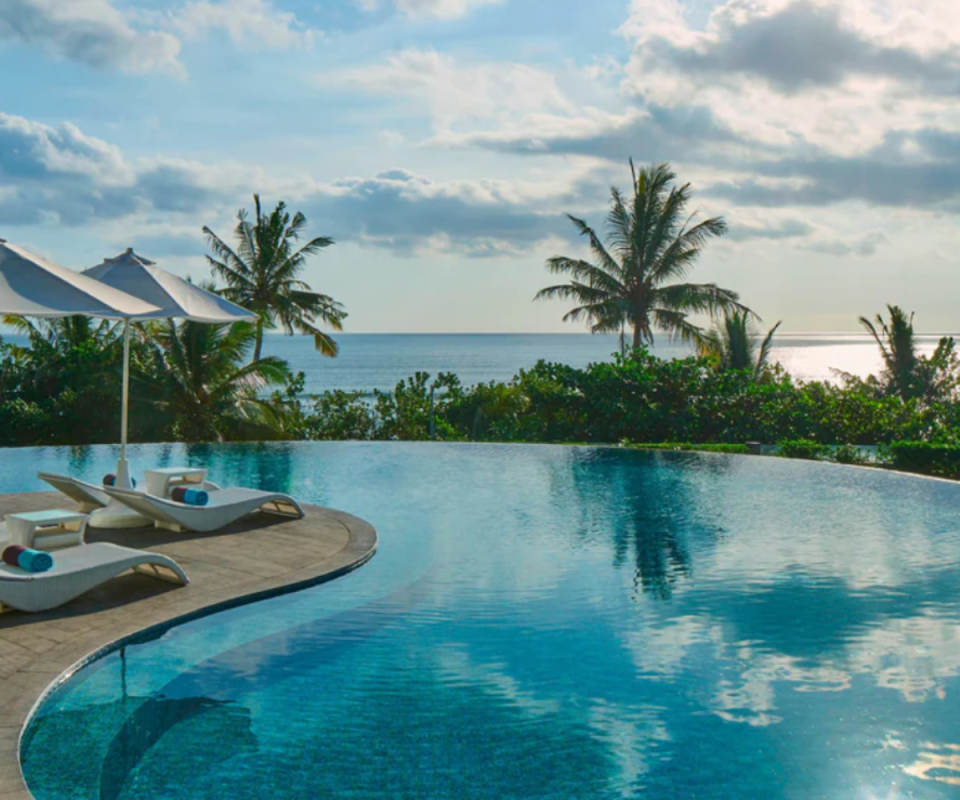 Looking over a swirling curved blue pool with a deck on the left with white deck chairs and umbrellas, with the ocean and palm trees in the background ahead. 