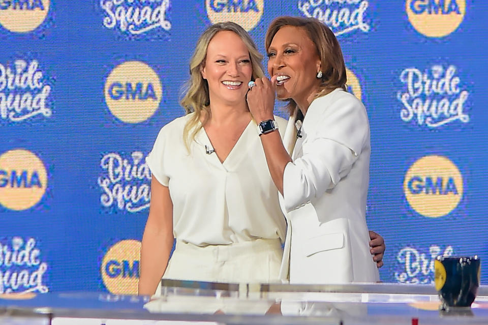 Amber Laign and Robin Roberts, dressed in white, smile and embrace on the GMA set, with a "Pride Squad" background