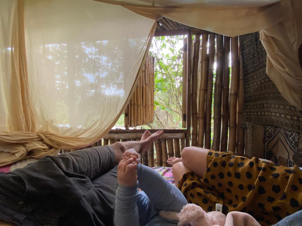 parents and a baby laying in bed with only their legs visible and curtains and window open.