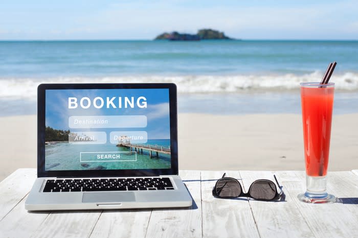 On a table at the beach, next to a tropical drink and a pair of sunglasses, a laptop shows a travel booking website.