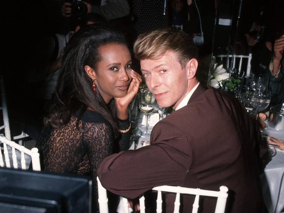 Iman in a brown dress leaning over to David in a brown suit sitting at a table.