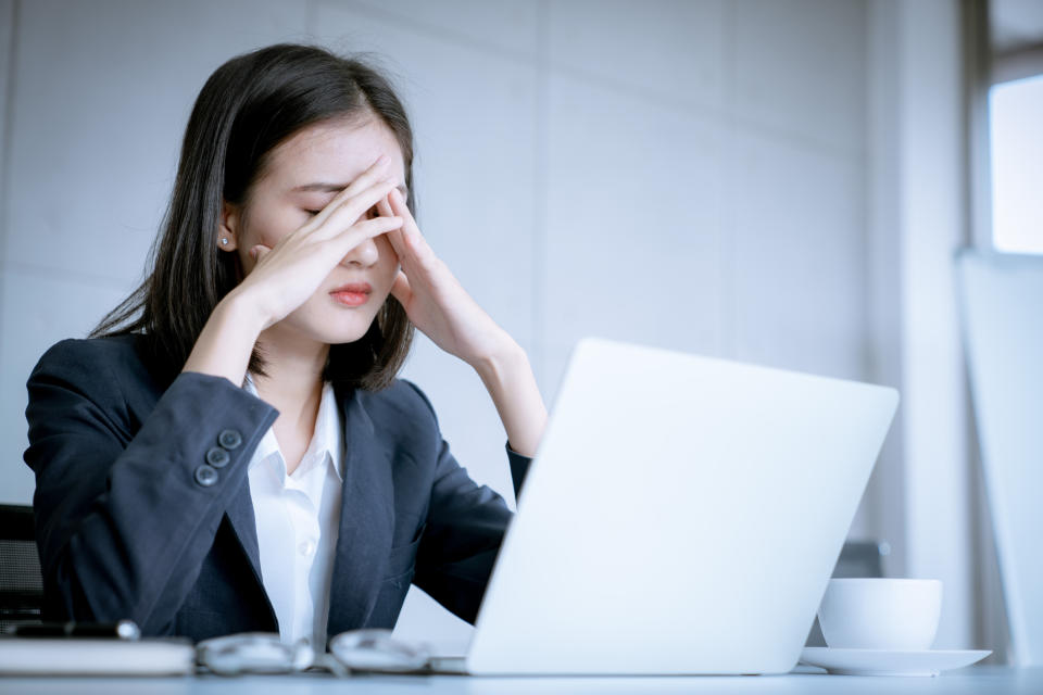 A woman in professional dress sits in front of a laptop, her hands covering her face in apparent exasperation.