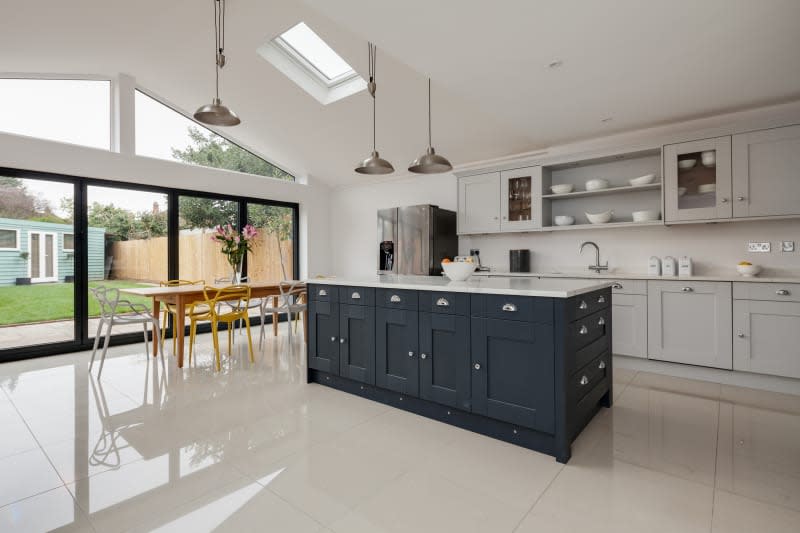 Contemporary modern luxury fitted kitchen with dark painted peninsular island unit, dining table with chairs and bifold patio doors leading to garden. High gloss floors