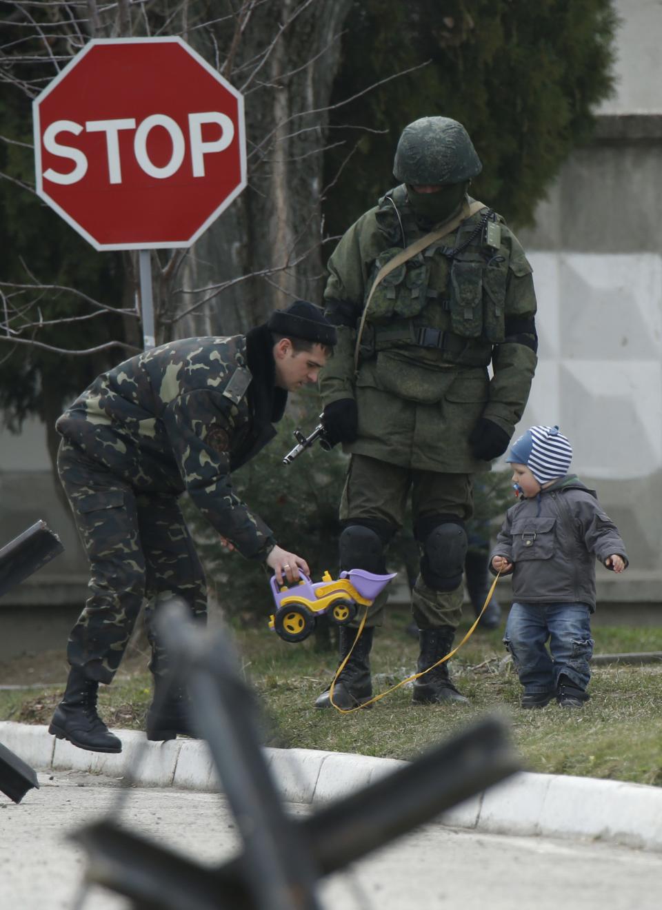 A Ukrainian serviceman plays with a child as a Russian serviceman stands in front of the gates of a Ukrainian military unit in the village of Perevalnoye outside Simferopol