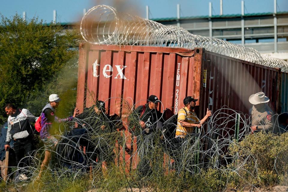 PHOTO: A Texas Department of Public Safety official, right, looks on as migrants walk near a rail car covered in Concertina wire at the Texas-Mexico border, on Jan. 3, 2024, in Eagle Pass, Texas. (Eric Gay/AP)