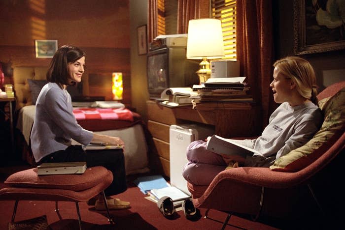 Selma Blair and Reese Witherspoon sit in a cozy room, smiling and engaged in conversation, while holding books in a scene from a movie